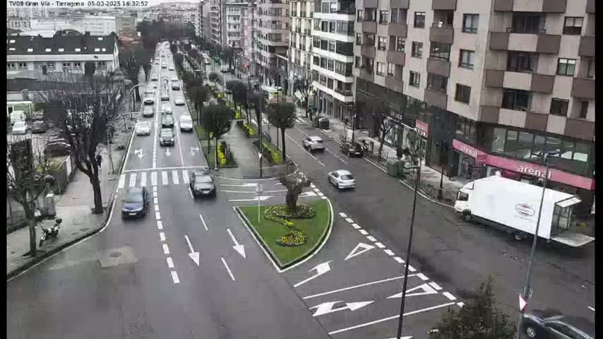 A city street scene on an overcast day shows several cars driving on a multi-lane road that intersects with a smaller road, and there are buildings lining both sides of the street.