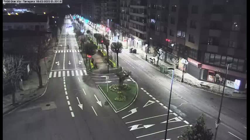 A nighttime, sparsely populated street scene shows a multi-lane road with streetlights and minimal traffic, bordered by buildings and landscaping.