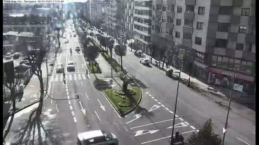 A sunny city street scene shows cars driving on a multi-lane road that intersects with a smaller road, with buildings and landscaping visible.