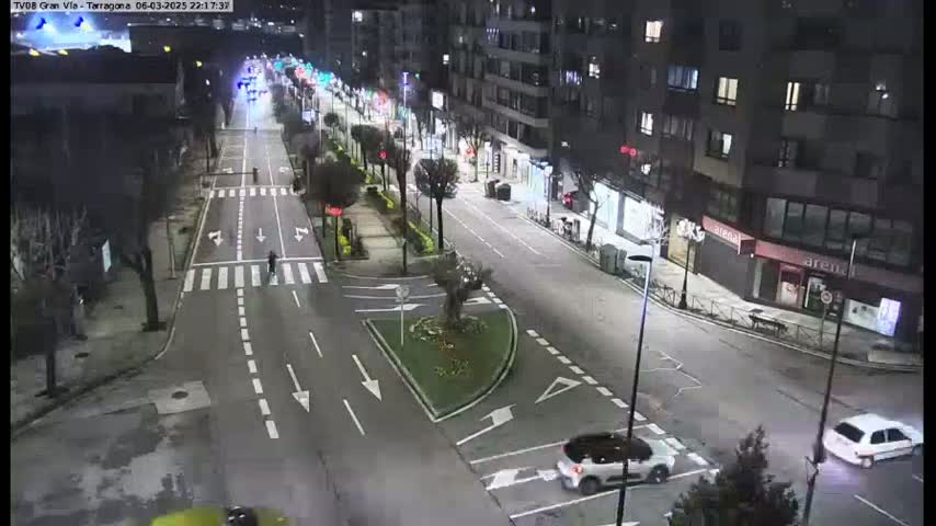 A nighttime, high-angle view shows a mostly empty city street with cars and pedestrians, and a small traffic circle in the center of the intersection.