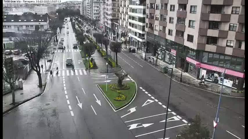 A wet city street scene shows several vehicles traveling along a multi-lane road lined with buildings and pedestrians crossing at a crosswalk, with some landscaping in a central median.