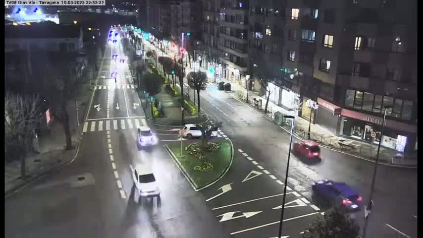 A nighttime, wet street scene shows several cars driving along a multi-lane road, with streetlights illuminating the area and a median with landscaping visible.