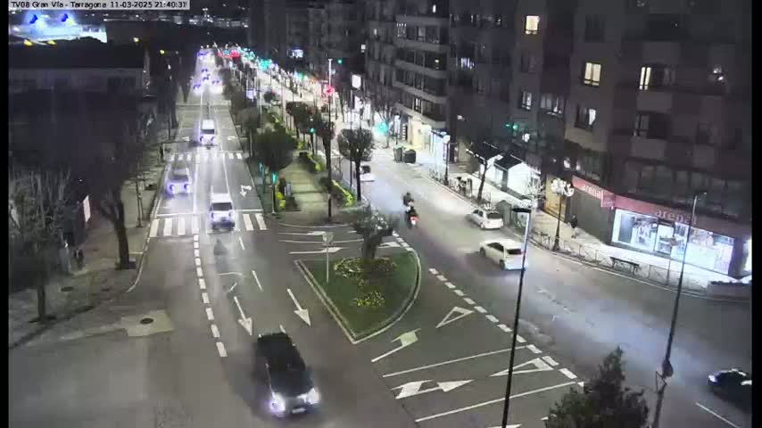 A nighttime, street-level view of a city intersection with several cars driving on the roads, lined with buildings and trees.