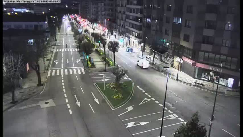 A nighttime, well-lit view of a city street intersection with sparse traffic, showing buildings and landscaping in the urban area.