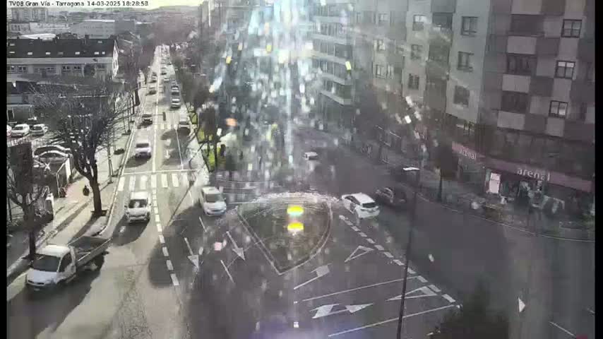 A sunny, slightly hazy day reveals a busy city intersection with several cars, a pickup truck, and buildings surrounding it.