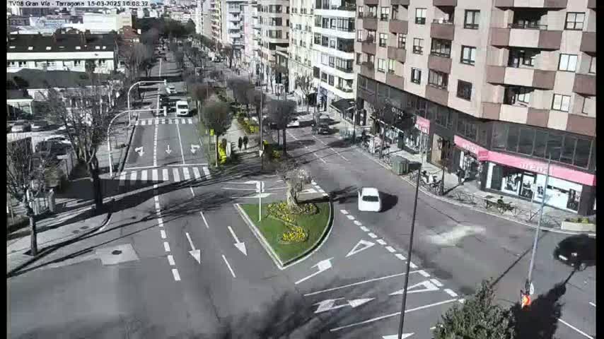 A sunny day shows a city intersection with several cars and pedestrians, a central traffic island with landscaping, and buildings lining the streets.