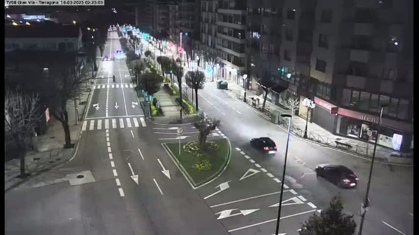 A nighttime, high-angle view of a mostly empty city street with several cars, streetlights, and buildings lining the road.
