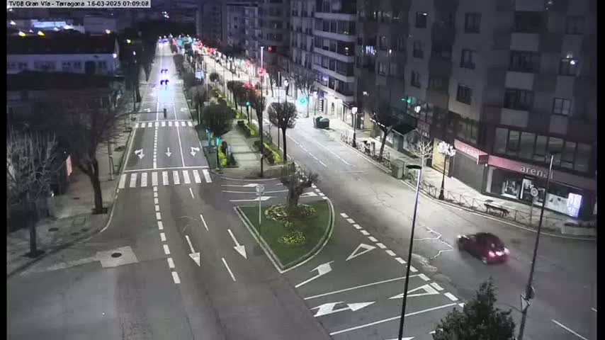 A nighttime, high-angle view of a mostly empty city street with a few cars, streetlights, and buildings lining the sides.