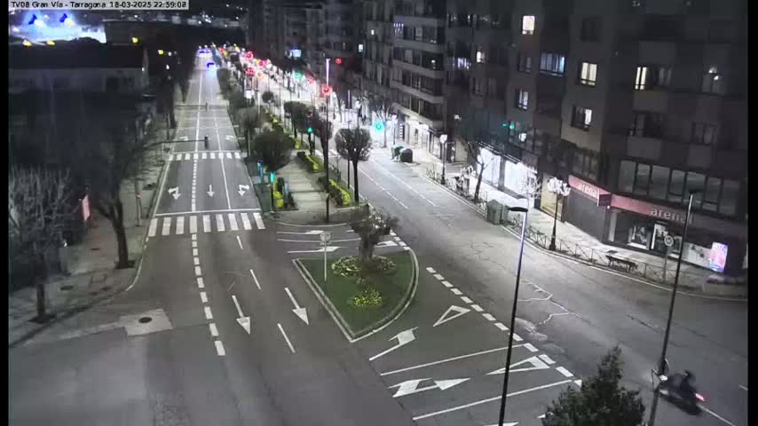 A mostly empty, well-lit city street at night, with a few cars and a motorcycle visible.
