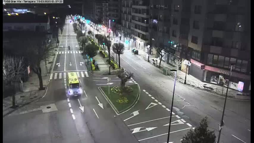 A mostly empty street at night, brightly lit by streetlights, with a single vehicle, possibly an ambulance, driving down the road.