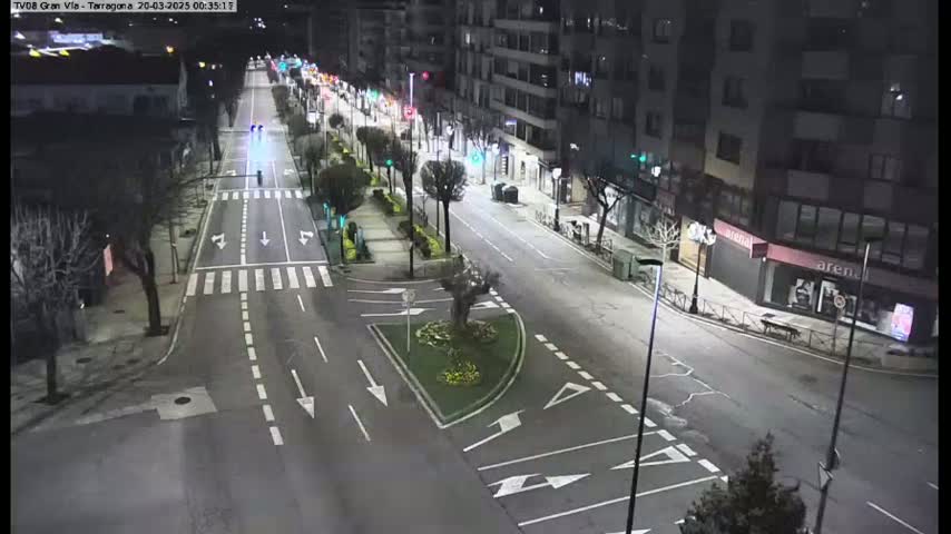 A mostly empty, well-lit street at night with a few pedestrians and vehicles, lined with buildings and trees.