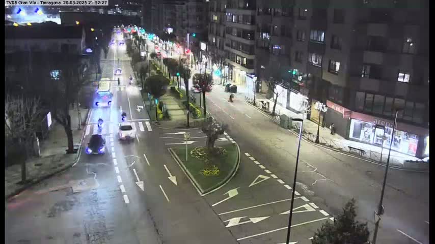 A nighttime, city street scene shows several vehicles driving on a multi-lane road, with pedestrians and motorcyclists also present, and the street is lined with trees and buildings.