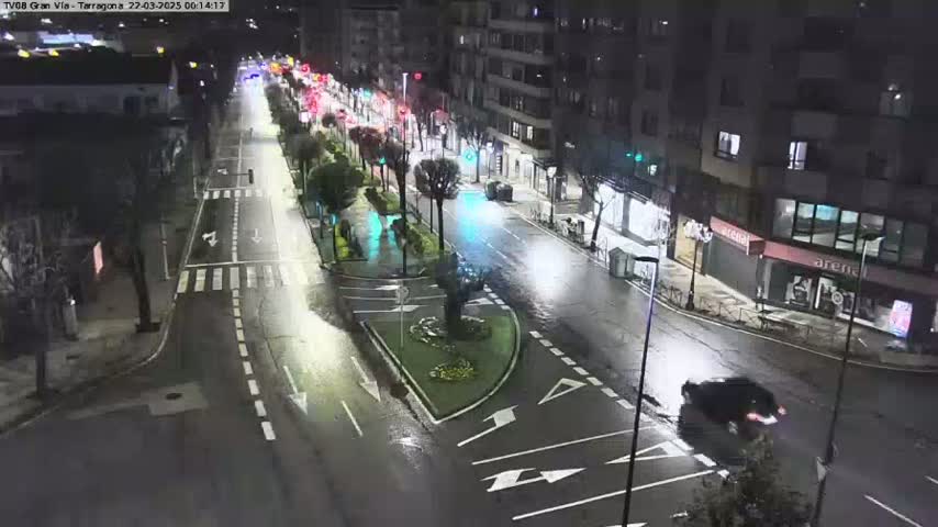 A wet, nighttime street scene shows a sparsely trafficked road with buildings lining both sides, and a few trees and small plants in landscaped areas.
