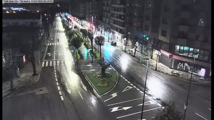 A wet, nighttime street scene shows a sparsely trafficked road intersecting with a busier avenue lined with buildings and trees under bright streetlights.