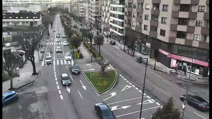 A city street scene with several cars driving on a multi-lane road, intersecting with another road, with buildings on both sides and a small landscaped area in the center, under a cloudy sky.