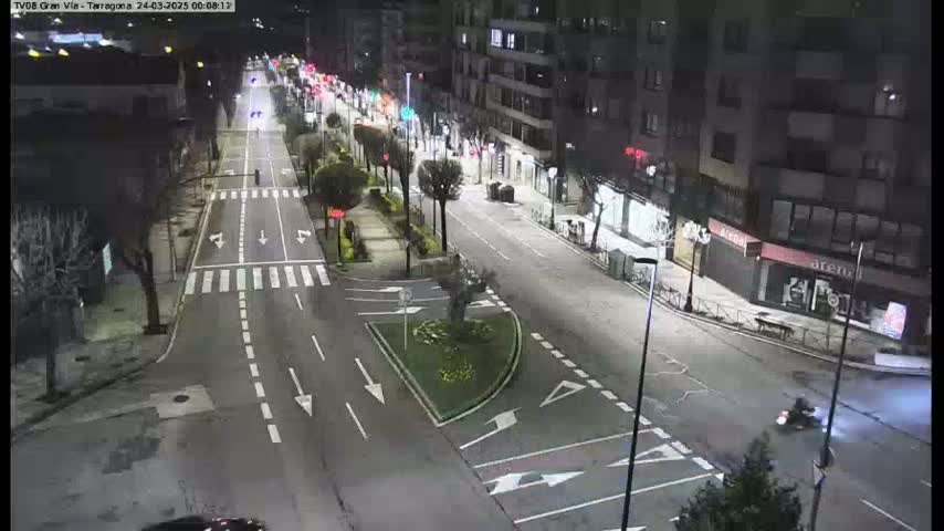 A nighttime aerial view of a city street with sparse traffic, illuminated by streetlights, and a small median with landscaping.