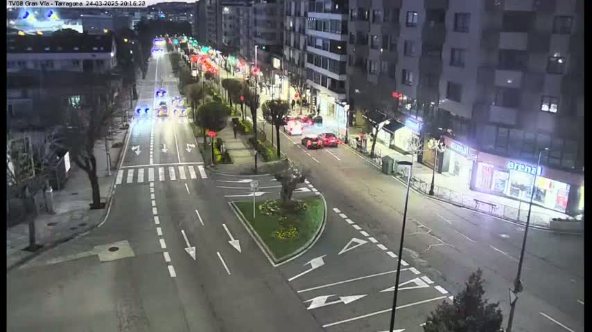 A nighttime, city street scene shows several cars driving along a multi-lane road with buildings and trees lining the sides.