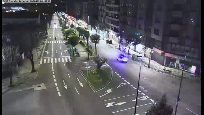 A nighttime, high-angle view of a mostly empty city street with a single car driving down the road, surrounded by buildings and streetlights.