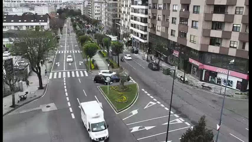 A mostly sunny day shows a city street intersection with several cars and a white van, surrounded by multi-story buildings and landscaped areas.