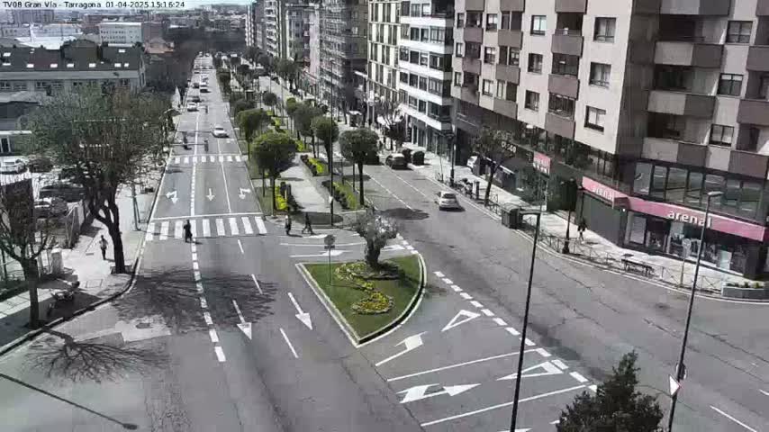 A sunny day reveals a city street scene with several cars and pedestrians navigating a multi-lane road lined with trees and buildings.