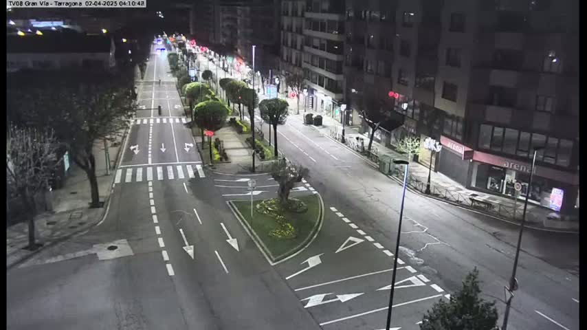 A nighttime aerial view of a mostly empty city street with some vehicles, lined with buildings and trees, under bright streetlights.