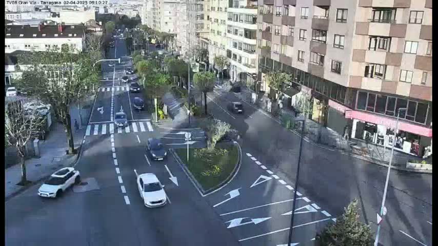 A partly sunny day reveals a high-angle view of a city intersection with several cars, buildings, and trees.