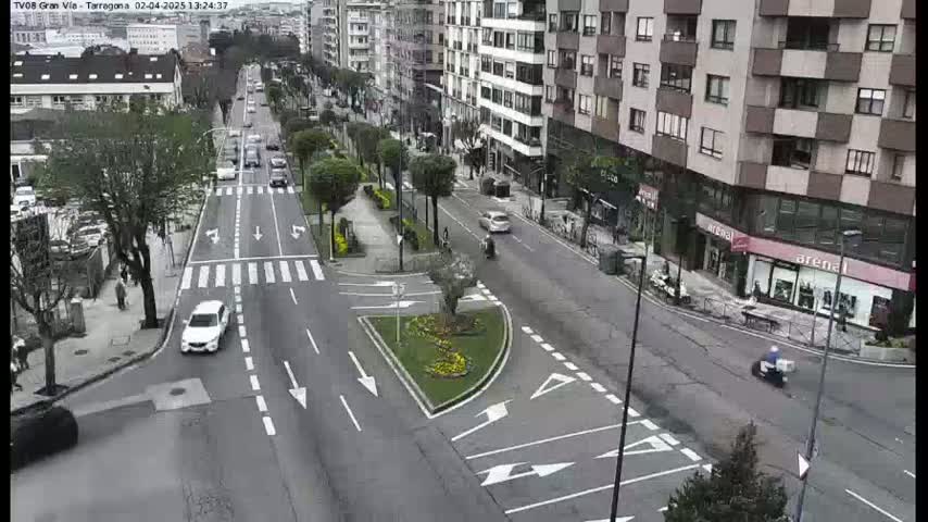 A city street scene on a cloudy day shows several cars driving on a multi-lane road that intersects with a cross street, with pedestrians and buildings lining the street.