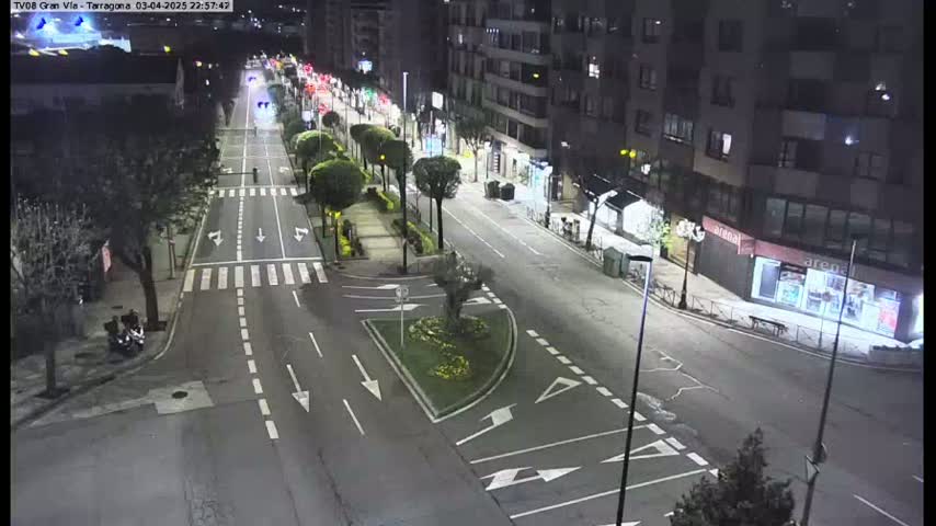 A nighttime street scene shows a mostly empty road with streetlights, lined with buildings and some landscaping, under clear skies.