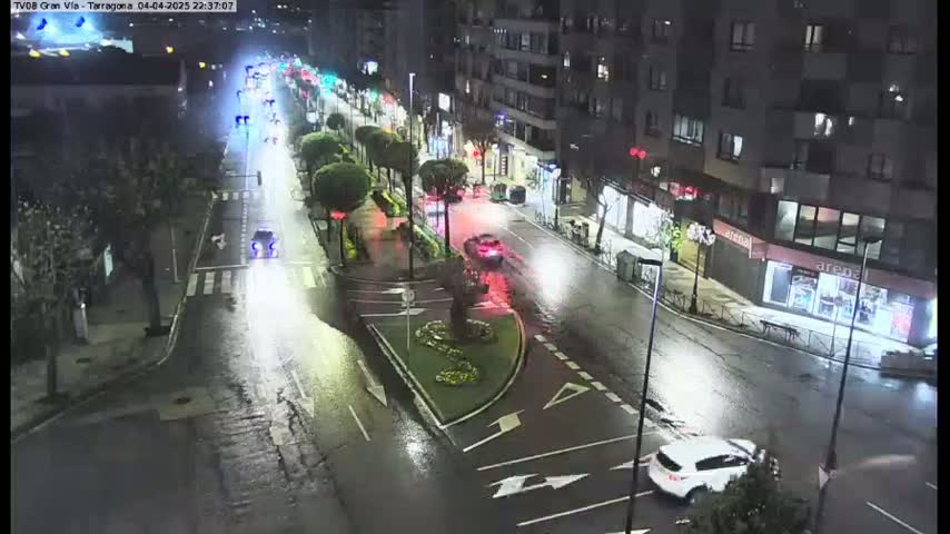 A wet, nighttime street scene shows several cars driving on a multi-lane road lined with trees and buildings.