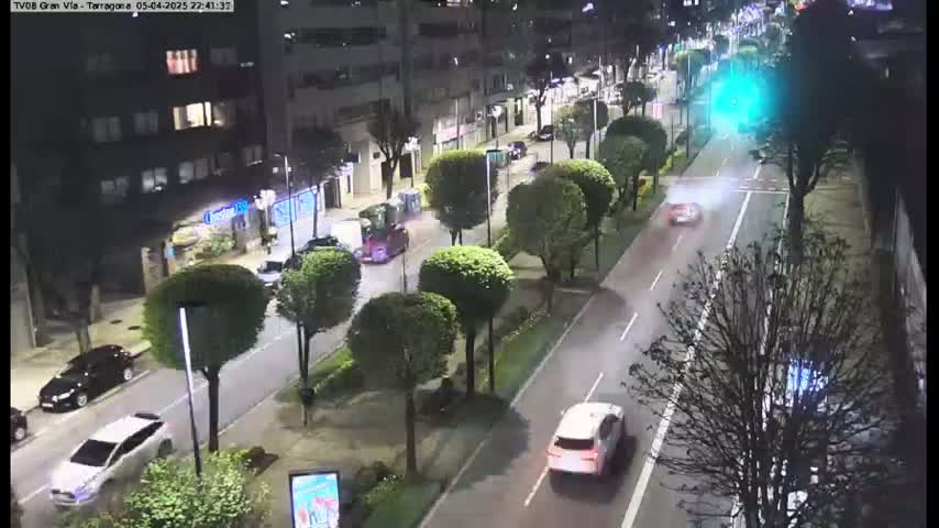 A nighttime, city street scene shows several cars driving on a multi-lane road lined with trees and buildings.