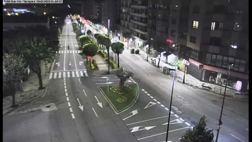 A nighttime aerial view of a mostly empty city street with buildings lining both sides, illuminated by streetlights.