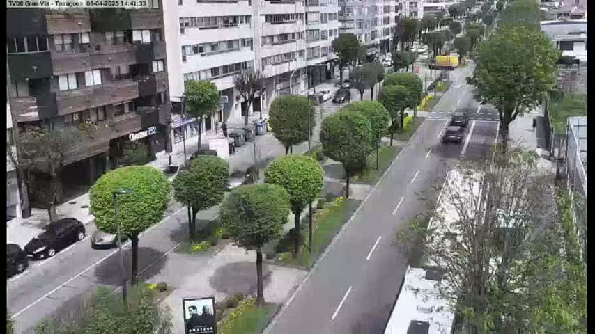 A paved street lined with green trees and parked cars runs past buildings under a partly cloudy sky.