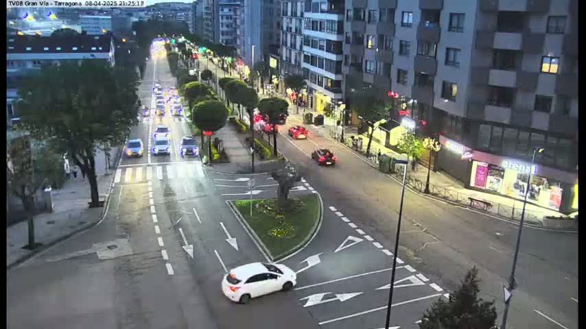 A white car is turning onto a street at night, with several other vehicles on a multi-lane road lined with buildings and trees under clear skies.