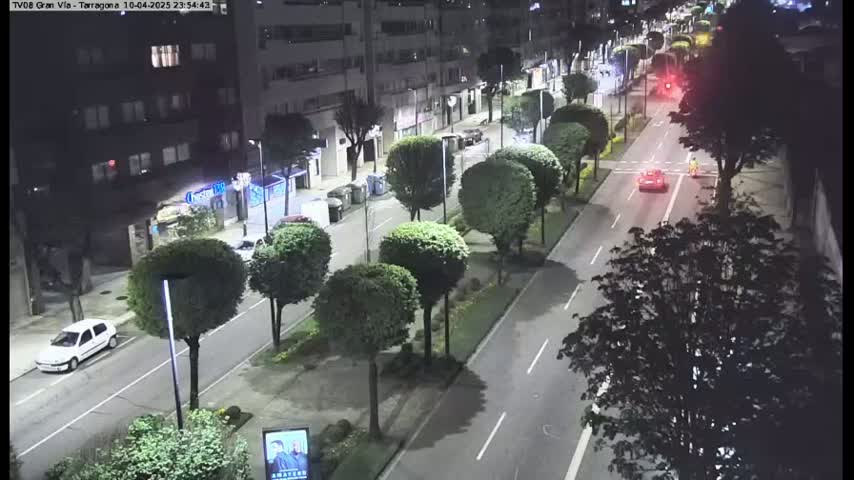 A nighttime, city street scene shows a few cars driving on a multi-lane road lined with trees and buildings, under well-lit conditions.
