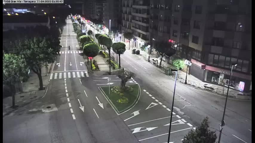 A mostly empty, well-lit street at night with buildings lining both sides, featuring trees and streetlights, and a small traffic circle in the center.