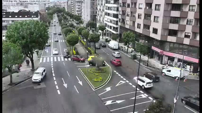 A city street scene shows several cars driving on a multi-lane road that intersects with another road, with buildings and trees lining the street on a partly cloudy day.
