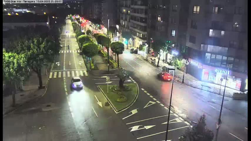 A wet, nighttime street scene shows several cars driving on a multi-lane road lined with trees and buildings.