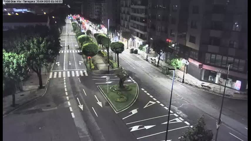 A nighttime, brightly lit street scene shows a mostly empty road with lane markings and a small, tree-centered traffic island.