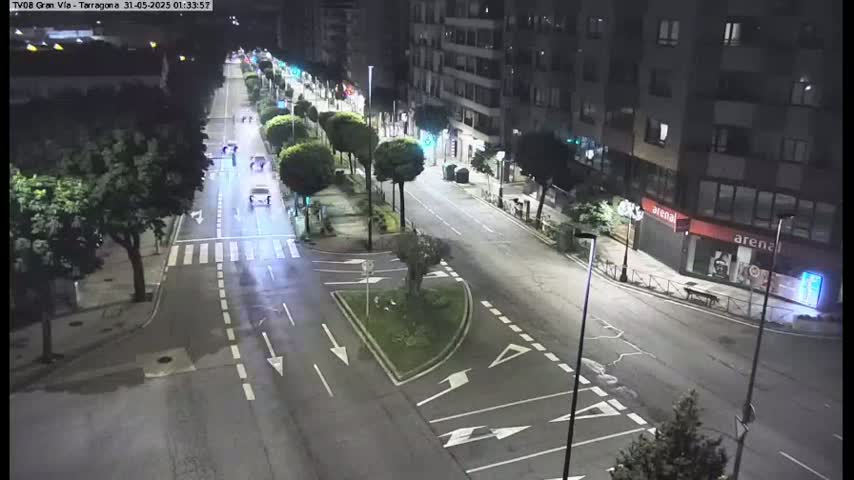 A nighttime, sparsely trafficked street scene shows a multi-lane road intersecting with another road, lined with trees and buildings.
