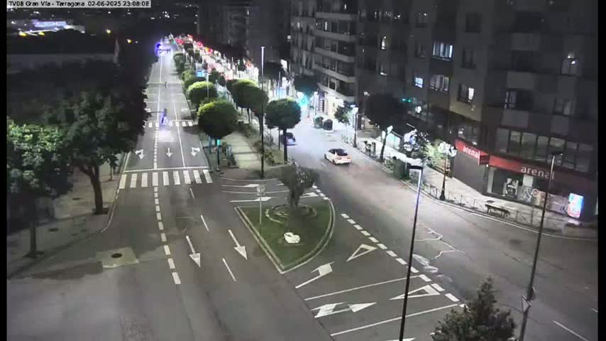 A nighttime, brightly lit city street scene shows a few cars driving down a wide road lined with trees and buildings.