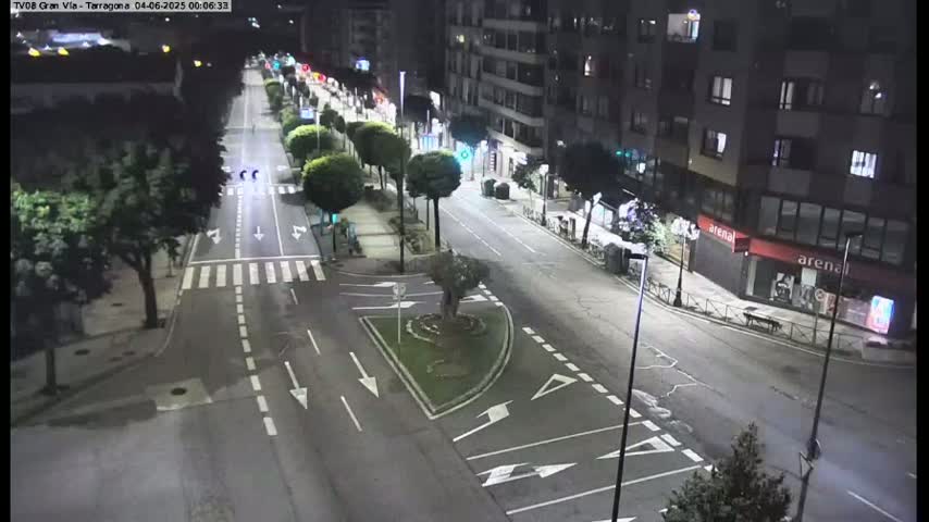 A nighttime, city street scene shows a mostly empty road with buildings on either side, lined with trees and streetlights.