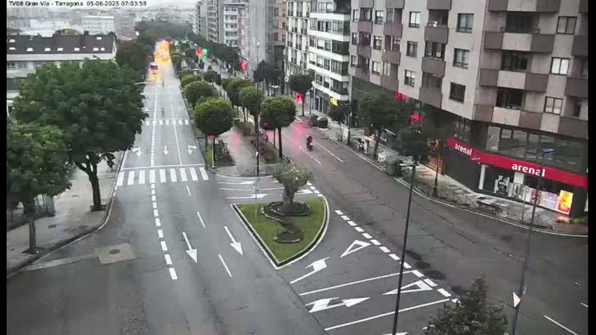 A wet, city street scene shows a multi-lane road with sparse traffic, lined with buildings and trees.
