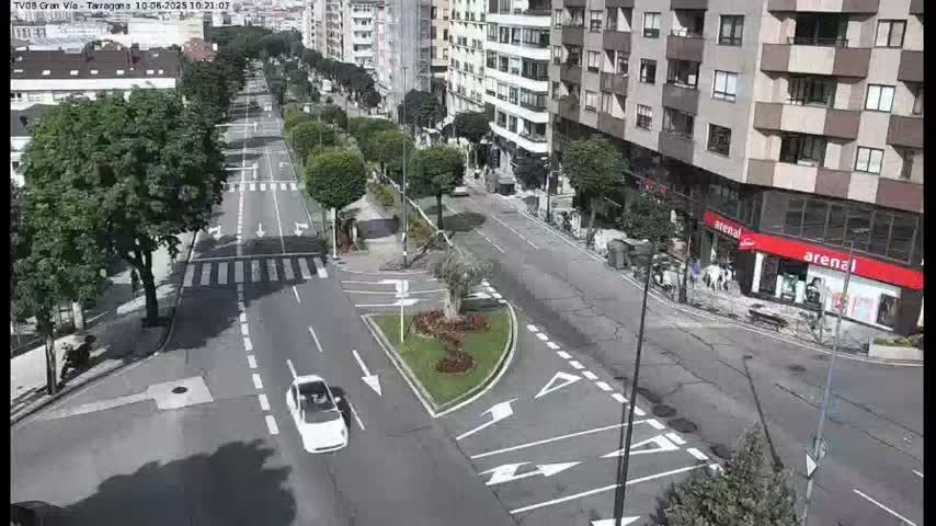 A mostly empty city street with buildings on either side, a small traffic circle in the center, and a white car driving down the street on a partly sunny day.