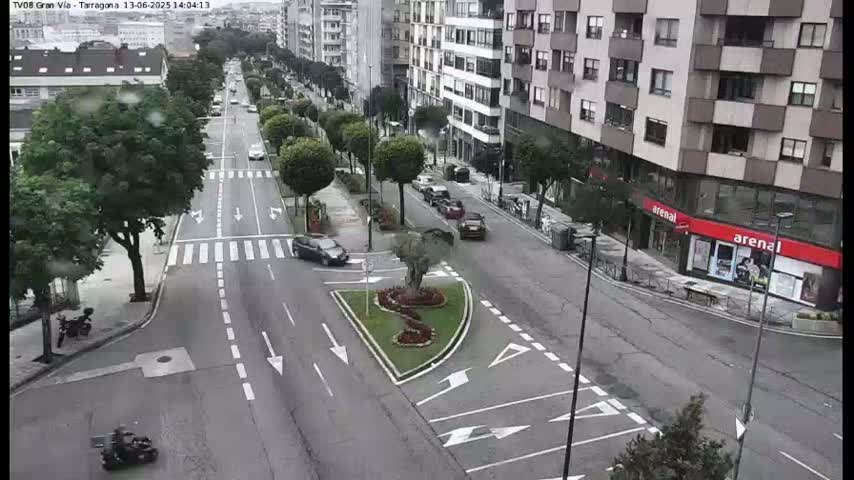 A city street scene on an overcast day shows several cars driving on a multi-lane road lined with trees and buildings.