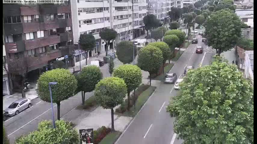 A city street lined with lush green trees has several cars driving down it on a partly cloudy day.