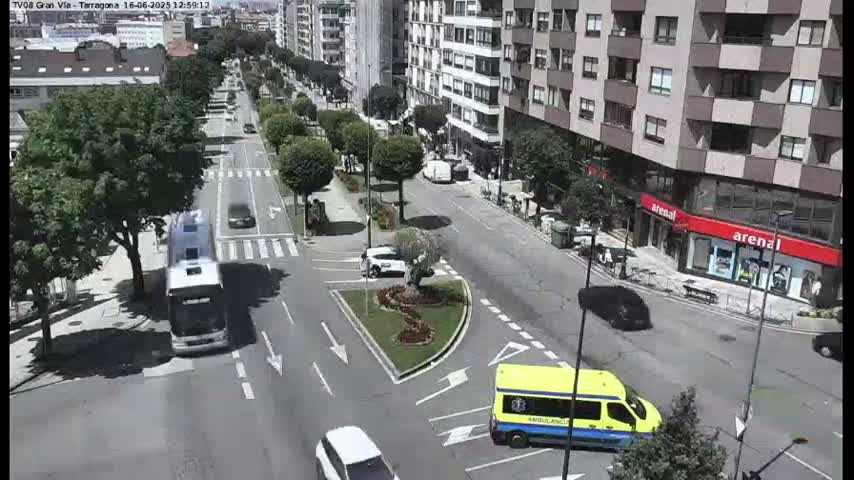 A sunny day shows a city street scene with several vehicles, including a bus and an ambulance, moving along tree-lined avenues and around a traffic circle.