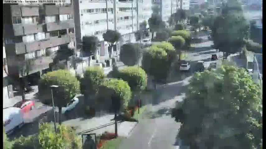 A street scene shows several cars driving on a road lined with apartment buildings and lush green trees under a partly sunny sky.