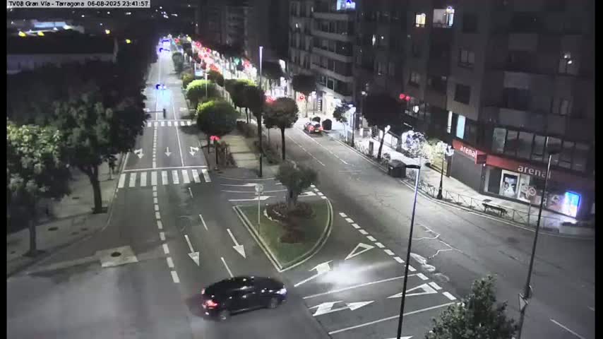 A nighttime, high-angle view shows a mostly empty multi-lane road with cars and streetlights, lined with trees and buildings.