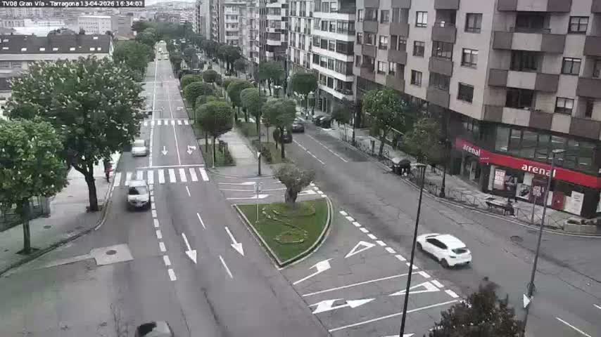 An elevated view captures a city street lined with multi-story buildings and trees, featuring cars on the road and a few pedestrians on the sidewalks, all under clear, sunny conditions.