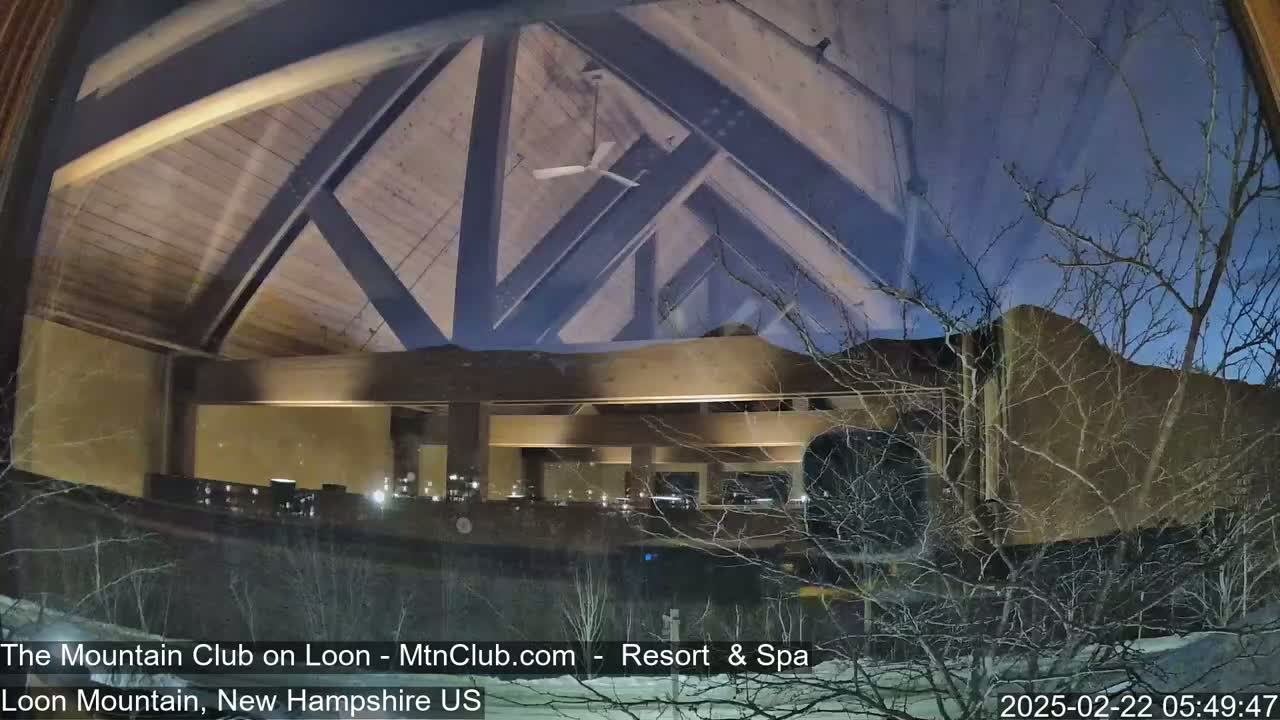 A nighttime view through a window shows a snow-covered landscape with bare branches and a building's interior with a wooden ceiling and warm lighting.